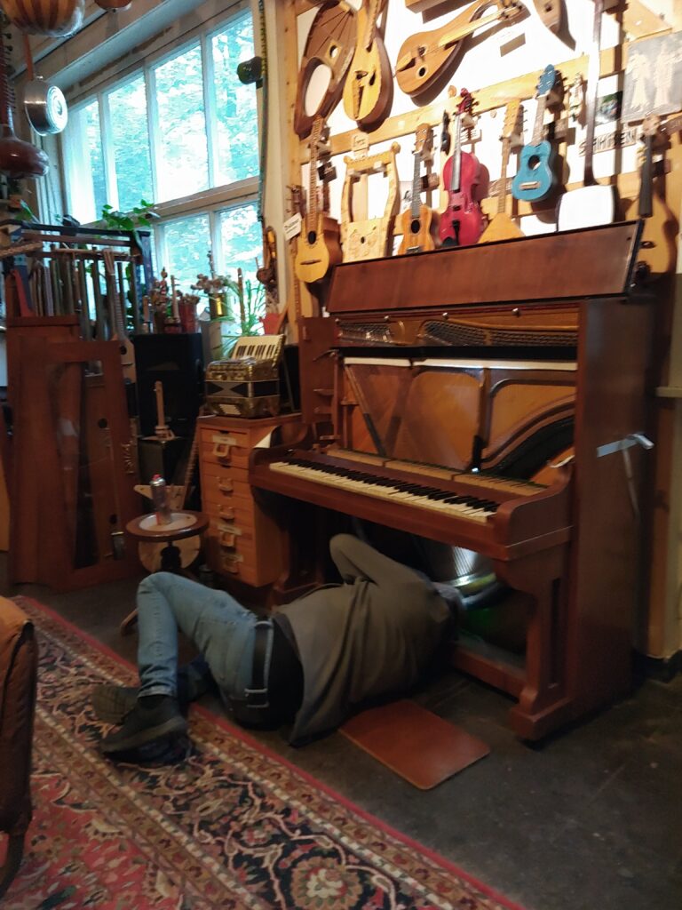 Das Klavier bekommt ein Licht - der Kasten ist aufgeklappt, die Saiten sind sichtbar und eine Person liegt auf dem Boden mit dem Kopf unter der Tastatur um die Lichtleiste festzumachen.